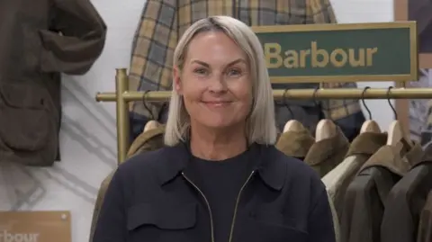 BBC Woman standing in the foreground, wearing a dark jacket and top, facing the camera inside a clothing retail display. Behind the person is a rail of waxed jackets on wooden hangers and a green sign reading “Barbour”. Additional jackets hang on the wall, including a checked garment. A plaque on the left displays the text “Barbour Re-Loved”. The setting appears to be a branded clothing shop or exhibition area.