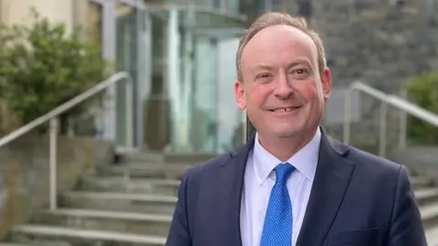 BBC Man in a suit with a blue tie standing in front of the royal court