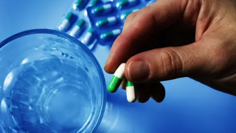 Fairfax Media A hand holding two white and green pills. There us a glass with some water in it. There are two blisters of pills on a blue table.
