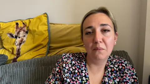 A woman is sat on a sofa. She looks sad. Behind her is a yellow cushion with a giraffe on it. 