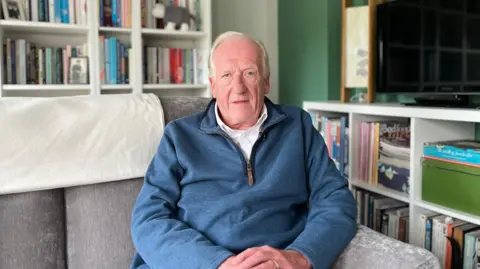 Gavin Walker sits on a grey sofa in a living room with a white bookcase behind him