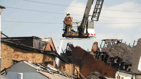 A firefighter is on an aerial platform ladder which is positioned over the top of a building. The building is black from fire and parts of the roof is missing, with exposed metal. The firefighter is blasting water at the roof with a hose