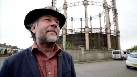Jonny Michel/BBC Joe Mackintosh is bearded and wearing a trilby hat. He is also wearing a blue jacket with a rust-brown shirt beneath. He is standing near the Great Yarmouth gasometer, which sits within a walled compound. There are vehicles parked on the right of the image, and a terrace of houses visible on the left of the image.