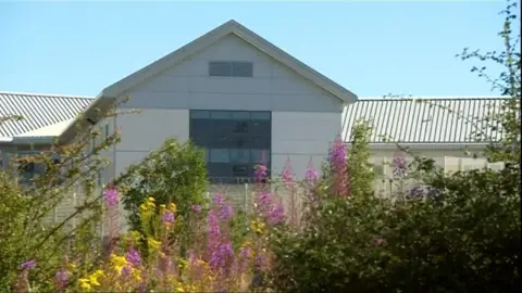 The outside of a prison. A beige building with a slatted roof. The middle section of the building has a large square window. Pink flowers and greenery surrounding the prison