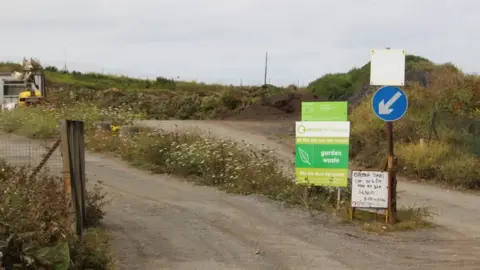 BBC The entrance of a garden waste centre. On the right are two signs. The one of the left is green and there is a smaller white one on the right. There is also a road sign above that. In the background is grass and flowers with a road at the centre.