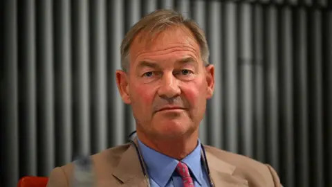 Getty Images Rupert Lowe in a tan suit, blue shirt and red tie wearing neutral expression against dark corrugated background