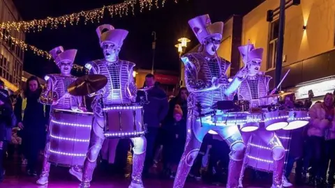 A music band with four members dressed in futuristic purple outfits performing in the street with lights on their instruments  