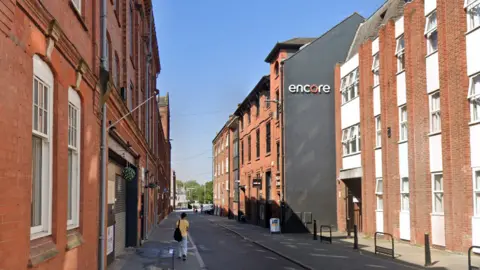 A street view of Millstone Lane in Leicester showing the Encore student flats building on the right and a person walking along the pavement on the left