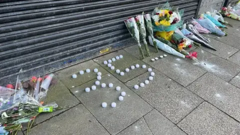 Floral tributes laid against the closed black shutters of Legacy Independent Funeral Directors. There are also some candles laid out in the shape of 35. 