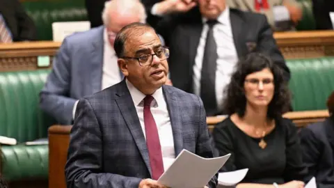 UK Parliament MP Mohammad Yasin speaking in the House of Commons regarding the closure of Bedford Renal Unit last year. He is wearing a checked suit, red tie and has black glasses on.