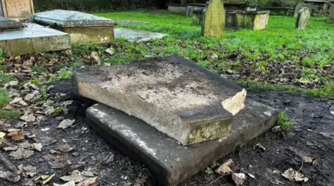 A broken gravestone, two large pieces of stone, one on the ground with mud and leaves around it, with the other piece of stone on top of the other.
