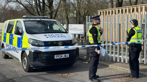 Two police officers stand behind a line of police tape beside a police van. In the background is an NHS sign saying "Hunter Street Health and Care Centre".