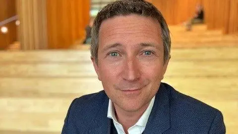A man sits on steps indoors, wearing a blazer and jeans. He has a slight smile and short, dark to grey hair.
