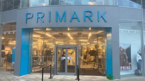 A glass fronted building, with large glazed panels and double doors, with temporary barriers outside the front. The building has the Primark brand marked in blue above the door, and blue and grey cladding on the pillars.