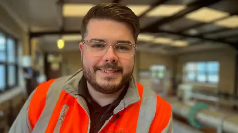 Fiona Irving / BBC A man with dark short hair, a beard and glasses smiles at the camera. He is wearing a high-vis orange jacket. He is standing in an industrial-looking room. Behind him you can see pipes.