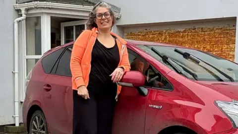 Nadine Lewis Nadine Lewis, who has wavy shoulder-length hair, standing next to a red car outside a house. She is wearing black-rimmed glasses and a bright orange fleece over a black top and trousers and has her hand on the car's wing mirror.