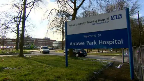 A blue and white NHS sign at the entrance to hospital grounds; the sign reads 'Welcome to Arrowe Park Hospital'.