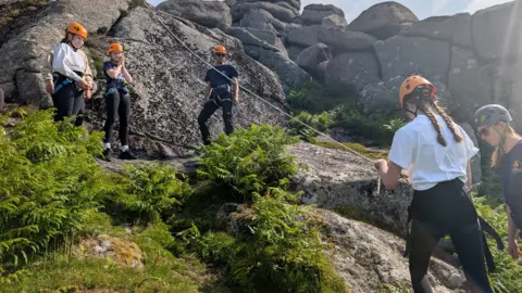 A girl with her back to the camera has just finished her abseil. Her friends stand at the top of the rope.