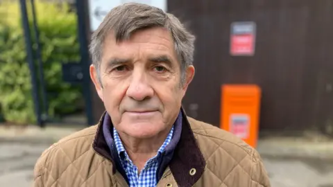 BBC Philip is wearing a brown quilted jacket and a gingham blue shirt. He has short brown and grey hair and has brown eyes. He is looking into the camera.