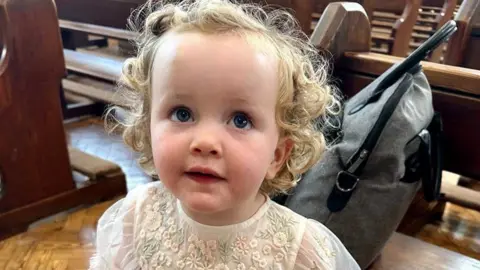 Family handout A small child in a white floral dress looks up beyond the camera at the person taking the picture. She has blonde curly hair, and appears to be standing in a church.