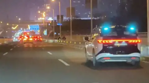 Reuters A view of a road at night time with a white and black car stopped in the middle of it - traffic continues further up the road.