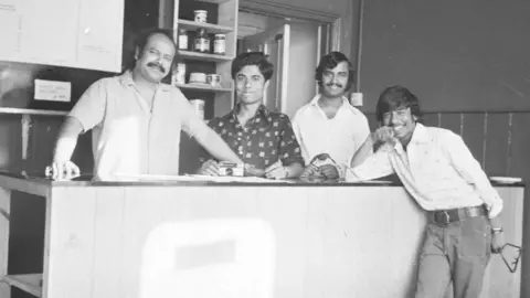 The Knights of the Raj Four people are looking at the camera, with three of them being behind what looks like a counter in the black-and-white image. Items on shelves and an open door are behind them.