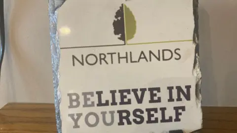 A small plaque sits on a wooden table. It says Northlands. Believe in yourself. An image of a leaf at the top.