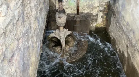 JOANNA TAYLOR/BBC Circular stone submerged by water with a a rod attached on top and brick walls on three sides. 