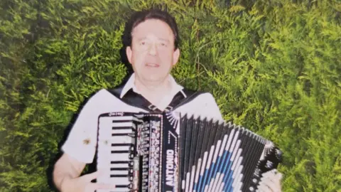 Family photo The late Ken Hopkins standing in front of a hedge, playing an accordion.  He has short, dark hair and is wearing a white, open-necked shirt. 