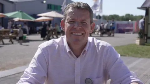 First Minister Rhun ap Iorwerth is sat down looking into the camera. He has short light brown hair, and is wearing a light, lilac shirt and is smiling. In the background there are festival-goers of the Urdd Eisteddfod, some are walking while others are sat on benches under parasols. There is also a large grey building far in the background and there are white flags or signs flapping in the wind.