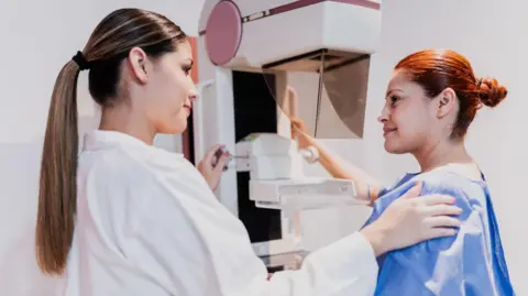 A female doctor doing mammogram exam on patient at hospital - stock photo. Both women have their hair tied back. The doctor is wearing a white lab coat and patient is wearing a blue gown.