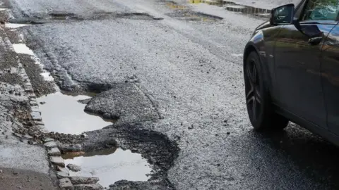 Two large potholes at the side of the road, with some more visible in the middle of the road a few yards ahead. A car can be seen just coming into frame on the right.