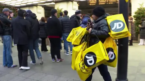 EVN / BBC Seorang pria dengan beberapa tas belanjaan dari JD sports berjalan melewati antrian Boxing Day di distrik perbelanjaan pusat kota London.