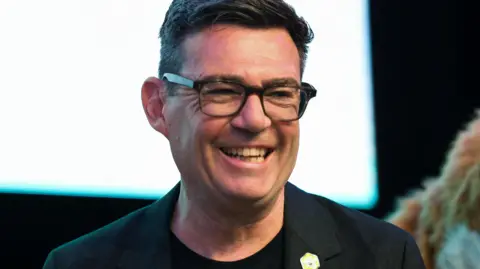 Newly elected Makerfield MP Andy Burnham smiles after winning the Makerfield by-election. He has short brown hair and is wearing a black blazer and t-shirt. He wears dark-framed glasses.