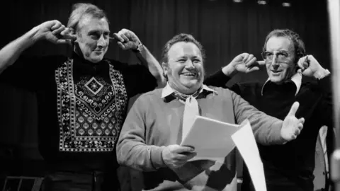 A black and white image of three smiling men, performing a show. The man on the left is tall, wearing a black jumper with an embroidered pattern. He has his fingers in his ears. The man in the middle holds a script is smiling, wearing a jumper, shirt and a tie. The man on the right also has his fingers in his ears, and wears large glasses, a dark jumper and a shirt with a large collar.