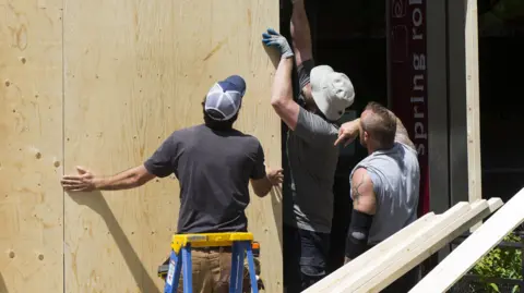 PA Media Three builders are looking at a large wooden panel in a shop window on a street. Two of them have their hands on it and the other has his arm on one of men's shoulders. The men wear T-shirts and hats and there are long lengths of wood in the foreground.