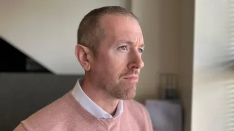 A man with short grey hair and stubble is standing in an office looking out a window which is out of frame. He is wearing a salmon jumper over a white shirt.