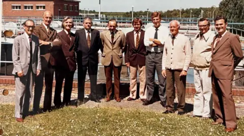Harwell Campus 10 men in a variety of 70s-era smart suits pose in front of a large industrial facility at Harwell Campus. It is a sunny day. Most of the men are aged between 40-70 and wear a variety of brown-coloured suits.