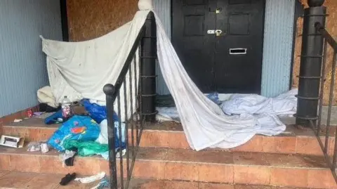 A doorway used by a rough sleeper. Steps lead up to a locked double door, with an entrance next to it blocked off by hardboard. Bedding is piled up at the top of the steps, with a sheet stretched across a railing and a post, and carrier bags and other litter strewn next to it.