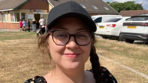Bryony Hill, a 40 year old woman wearing a black cap with plaited hair. Bryony, who has been diagnosed with an incurable brain tumour is a white woman, she has olive-toned skin and is wearing glasses with a square tortoiseshell frame. A community hall and car park can be seen in the background of the image. 