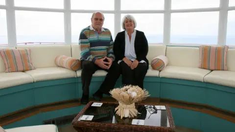 Simon Furber/BBC A man and woman sit on a cream sofa.