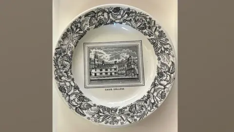 Caius College A white plate is decorated with a view of Caius Court in its centre, in black, and the words "Caius College" printed beneath. Around the edges are black drawings of flowers and foliage