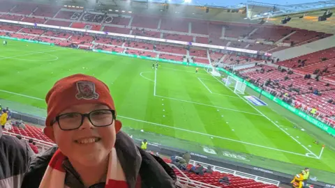 Family handout Riley is in a stand at the Riverside Stadium. He is smiling and has his back to the pitch where a few groundstaff are working. The stand on the opposite side and behind one of the goals can be partially seen. There are few fans in as it appears to be well before kick off. Riley is wearing a red Middlesbrough beany hat and red and white striped scarf. He is wearing rectangular black-rimmed glasses and a black coat.