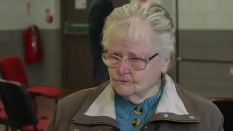 Esther Eastwood looks off camera and is wearing a brown coat with a blue cardigan underneath. She is wearing glasses and has short grey hair.