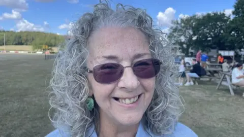 Lynne Bate has shoulder length curly grey hair and is wearing a blue top. She is also wearing oval-shaped sunglasses and is smiling at the camera. 