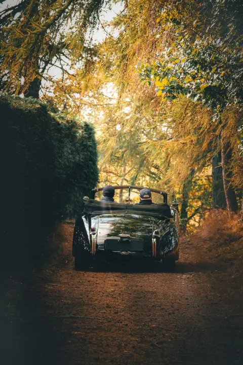 David Clem Vintage convertible car driving away down a narrow country lane, covered by trees, with two passengers in the car. The licence plate has been blurred.