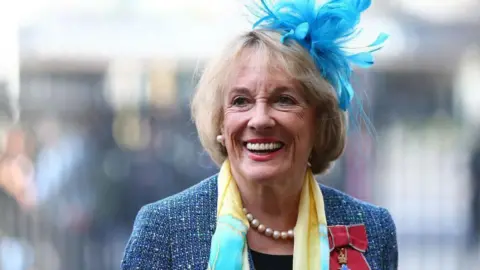 Getty Images Dame Esther Rantzen looks slightly off camera, smiling. She has short blonde hair with a fringe framing her face and wears large pearl stud earrings and a single row of pearls as a necklace to match. She is wearing a blue feather fascinator on her head, with a blue tweed jacket and a yellow and pale blue silk scarf around her neck and black top underneath. She has a red bow with a medal pinned to the left panel of her jacket - a symbol of her damehood.