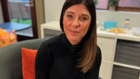 A woman with long dark hair and blue eyes looks into the camera. She is wearing a black jumper. Behind her is an orange cushion on a grey chair. There is a worktop behind the chairas well as a table and glass fronted door.