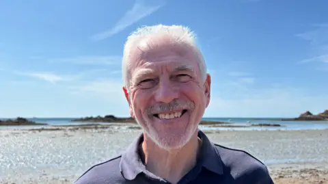 Kevin McIlwee is standing on a beach at low tide, wearing a dark navy polo shirt. The foreground consists of sand and shallow water reflecting sunlight, while rocky outcrops and small islets are visible along the horizon. The sky is clear and bright blue with thin wisps of cloud, creating strong natural daylight. The coastal setting is calm and open, with expansive sea views stretching behind him.