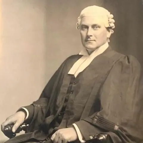 David Duggleby A black-and-white photo of a man dressed in a formal court gown and wig. He is sitting in a chair and looking towards the camera.